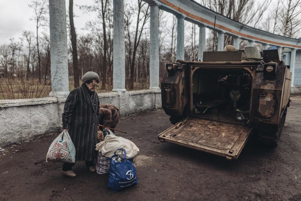 A woman in a fur coat with a walking stick and bags beside an armored vehicle