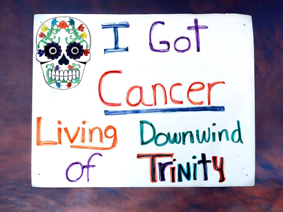 white paper sign with colorful skull illustrated and colorful words reading "I got cancer living downwind of Trinity"