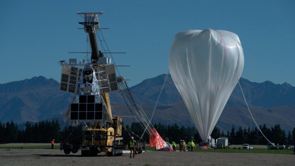  A large white partially inflated balloon rests on the ground, with mountains in the background. 