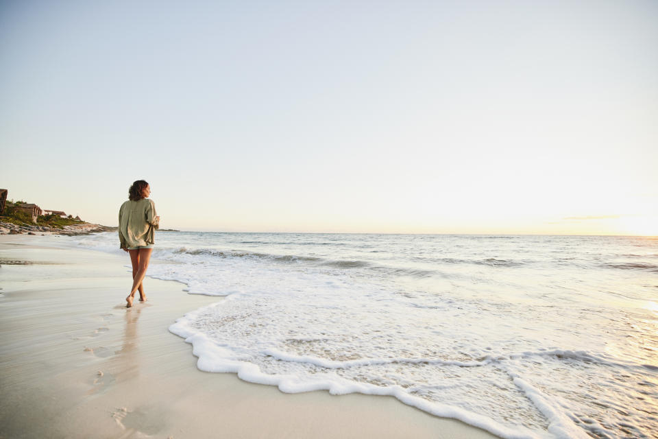 A person walks alone on a beach at sunset, leaving footprints in the sand, with waves gently washing onto the shore