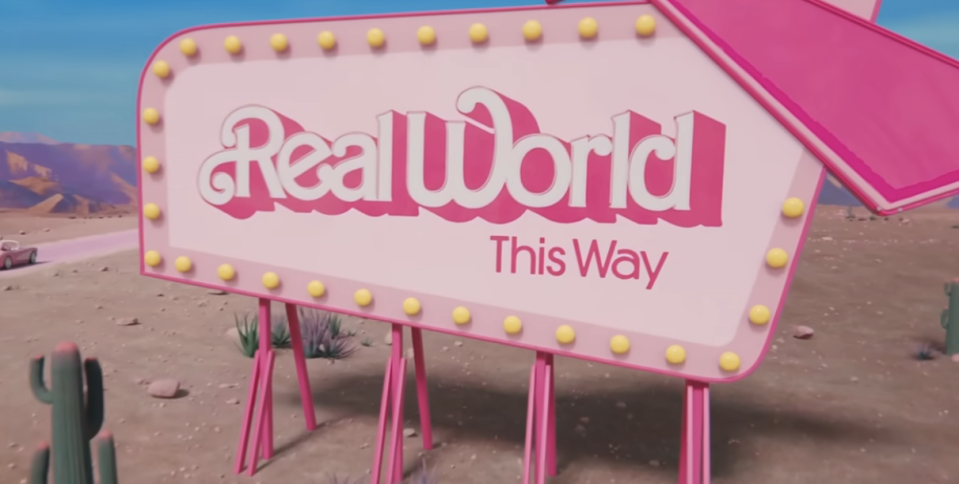 A sign by the road that says "Real World This Way" in a scene from Barbie