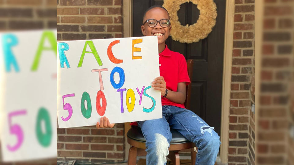 Orion Jean sits on a stool holding a sign that reads: Race to 500 toys.
