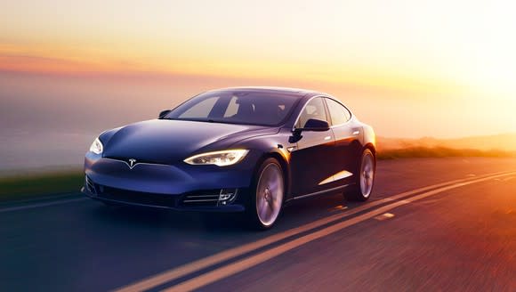 A Tesla Model S driving on a road along a body of water at dawn or dusk.