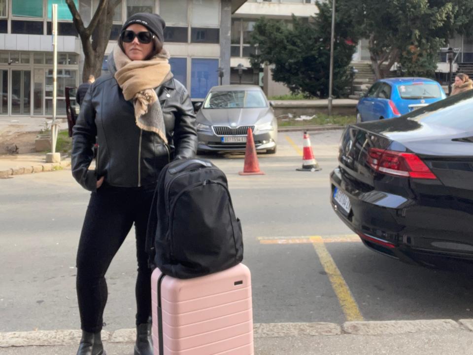 Katka, wearing sunglasses, a beanie, a chunky scarf, a leather jacket, boots, and black pants, stands on a sidewalk with a pink carry-on bag and a black backpack.