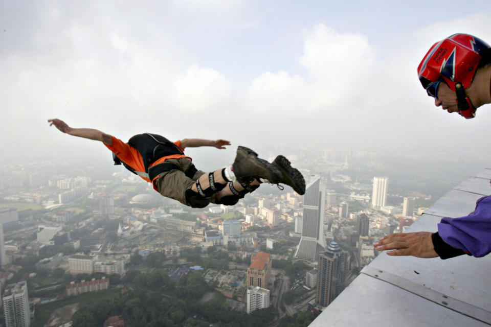 Base jumper in mid-leap from a high building, while another person watches. Cityscape in the background