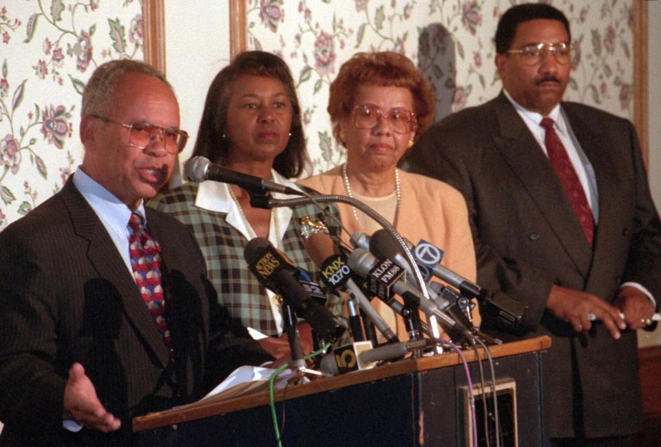 A man speaks at a lectern as three people stand to his left.