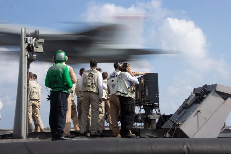 A group of sailors in vests observe a fighter jet landing