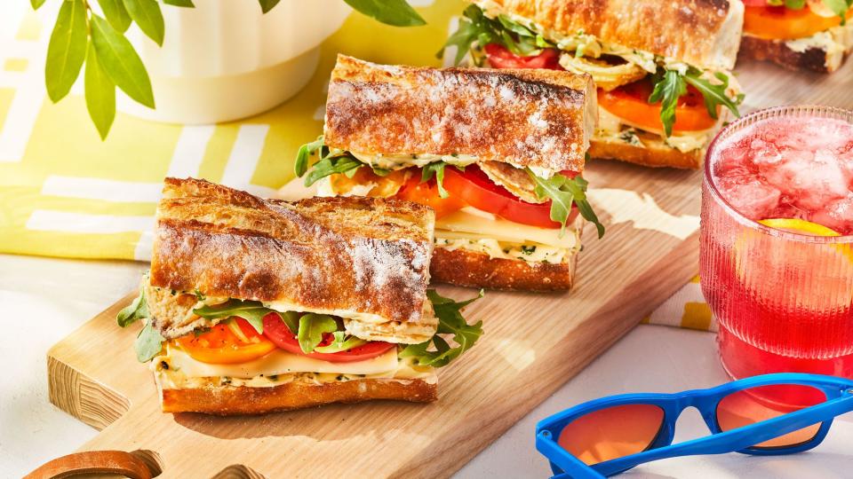 Fancy tomato sandwiches on a wooden board displayed on a table with fruit juice and sunglasses.