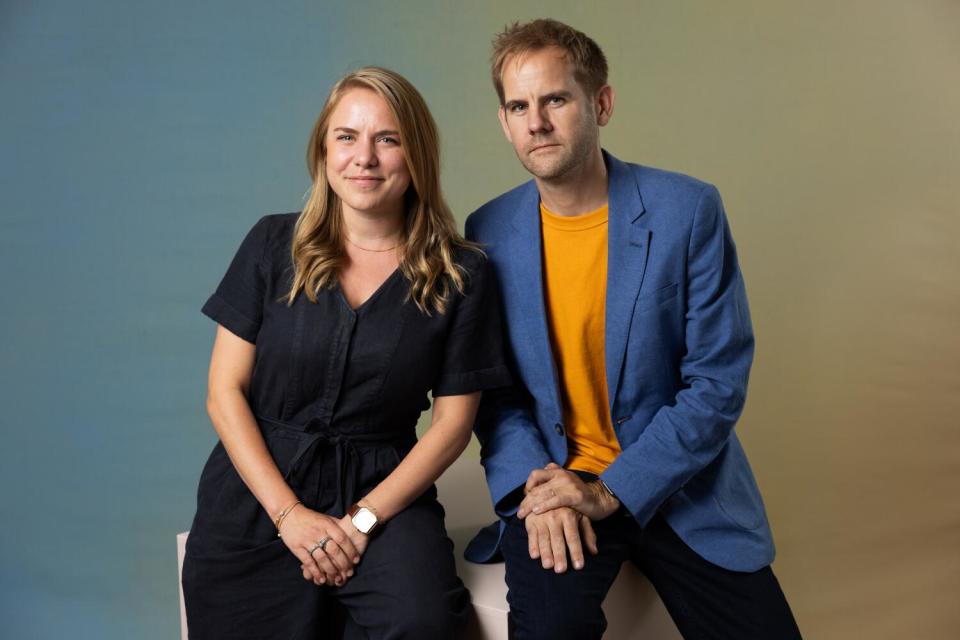 Co-directors Rachel Ramsay in a dark dress and James Erskine in an orange shirt and blue jacket sit on a cube