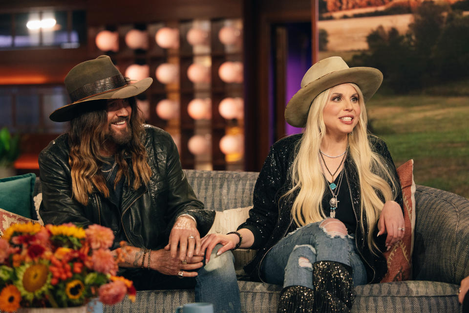 Billy Ray Cyrus and Firerose sit on a couch during a televised interview. Billy Ray wears a leather jacket and hat; Firerose wears a sparkly jacket and hat