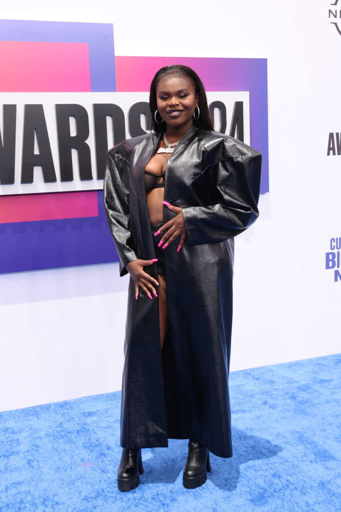 Scar Lip in a black, long, leather coat and matching boots posing on a blue carpet at an awards event. Background shows part of an awards sign