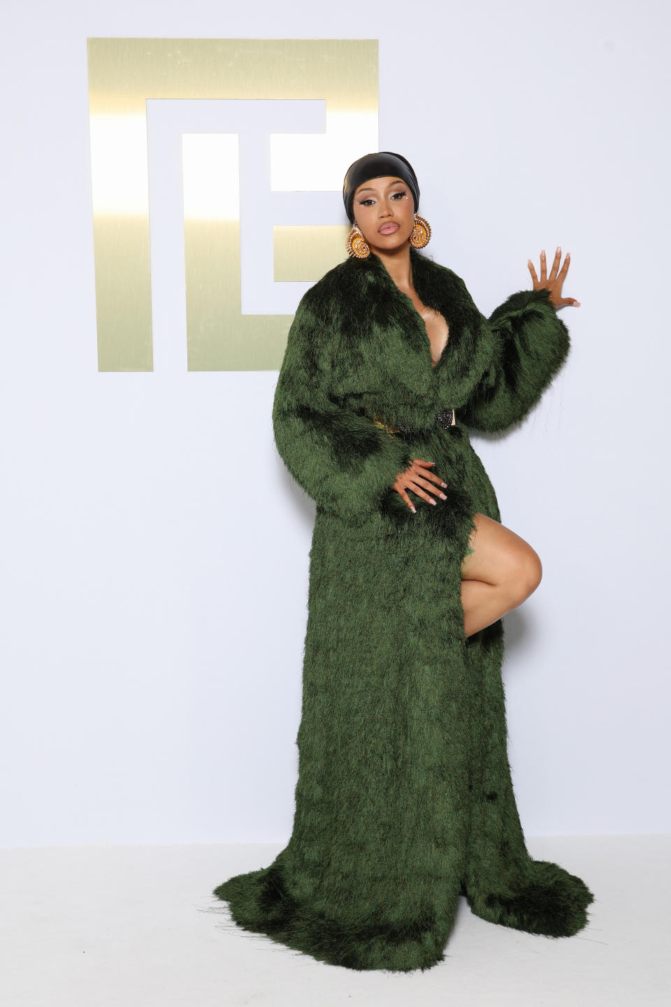 Cardi B poses in a long, fur-textured coat with a deep slit, gold statement earrings, and a headwrap, standing in front of an abstract, gold rectangle logo