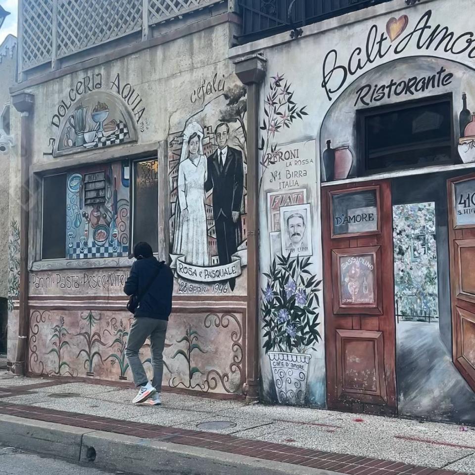 Person passing by mural-adorned building with balcony, possibly a restaurant, in a travel-related setting