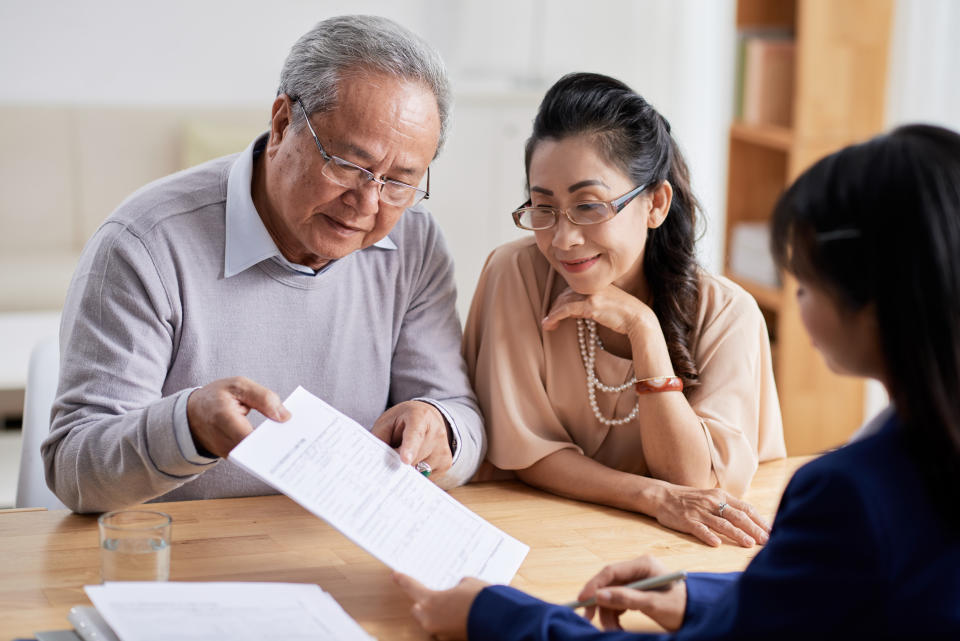 Concentrated senior man and his pretty wife studying terms of real estate purchase agreement while having meeting with realtor, interior of modern office on background