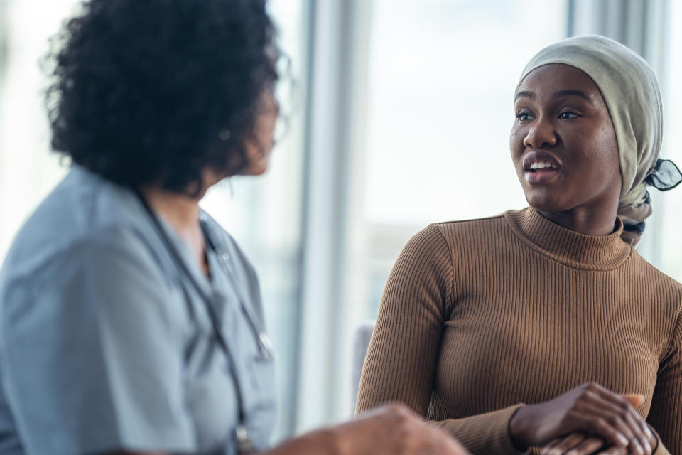 A young black woman with cancer is consulting her doctor. She is wearing a bandana to hide her hair loss. The two individuals are seated at a table together. They are discussing the patient's treatment plan.