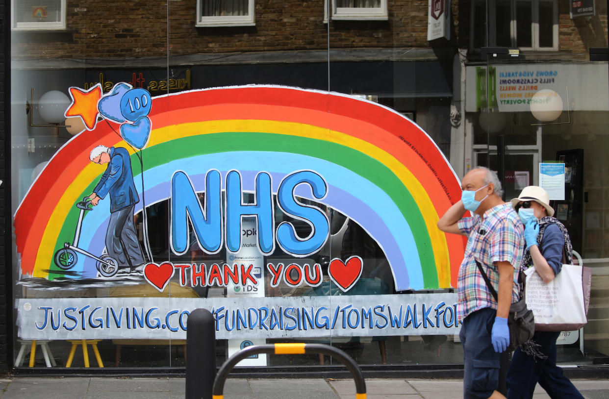 A couple wearing protective face mask and gloves walk past a window shop in Crouch End, north London, displaying an artwork depicting Captain Tom and a NHS rainbow to thank key workers for their efforts during the coronavirus pandemic. Picture date: Sunday July 12, 2020.