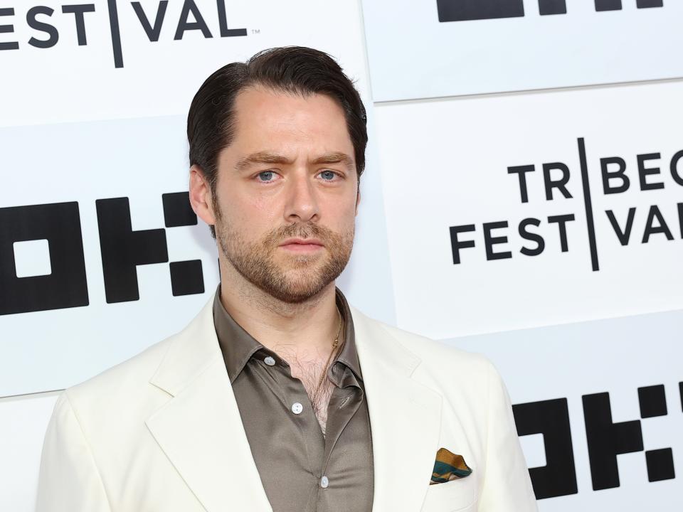 Richard Rankin wearing a white suit and a grey silk shirt.