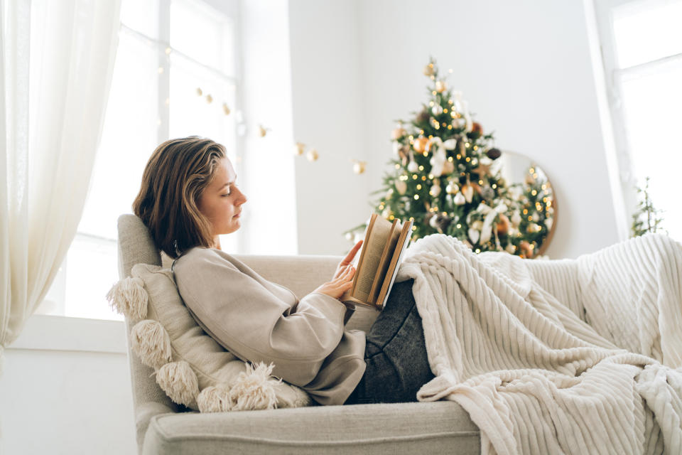 Young woman is reading book sitting on sofa near decorated Christmas tree with many lights. Concept of leisure during New Year holidays. Front view