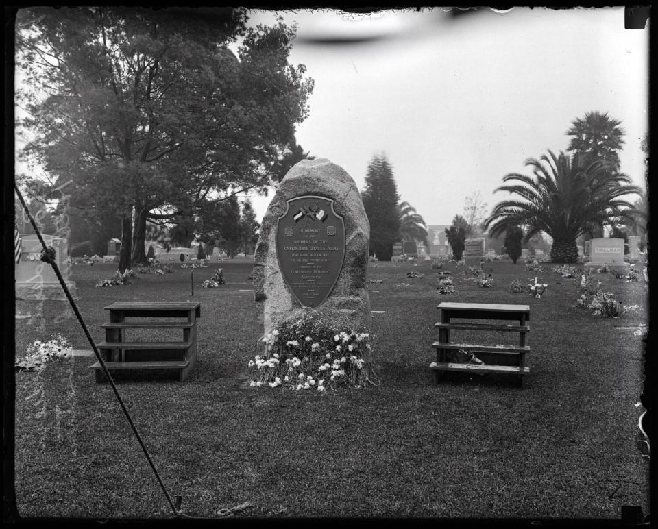 In a black-and-white photo with some damage, flowers rest at the foot of a stone monument with plaque in a cemetery.