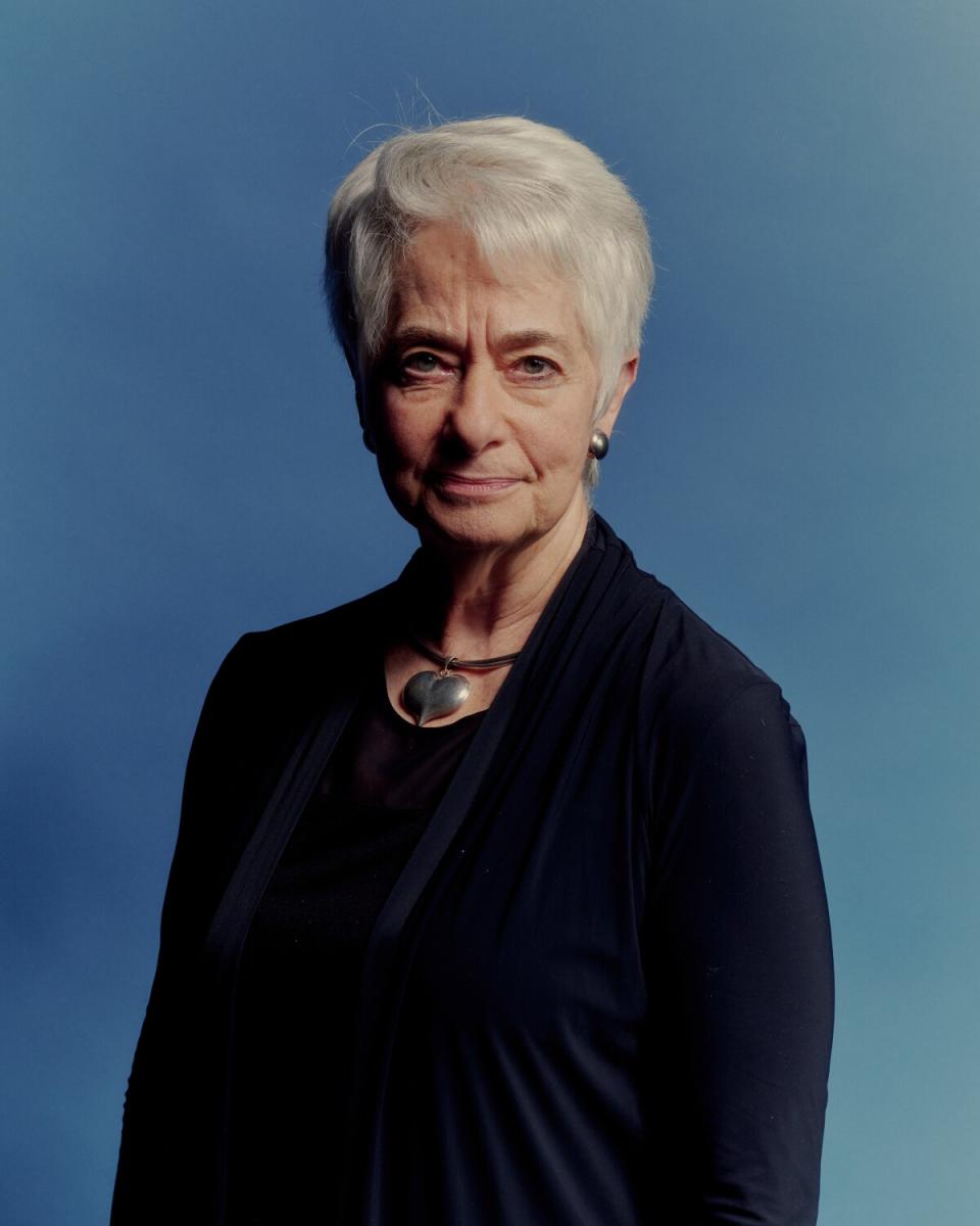 A woman in a black sweater and wearing a silver heart necklace stands against a blue background.