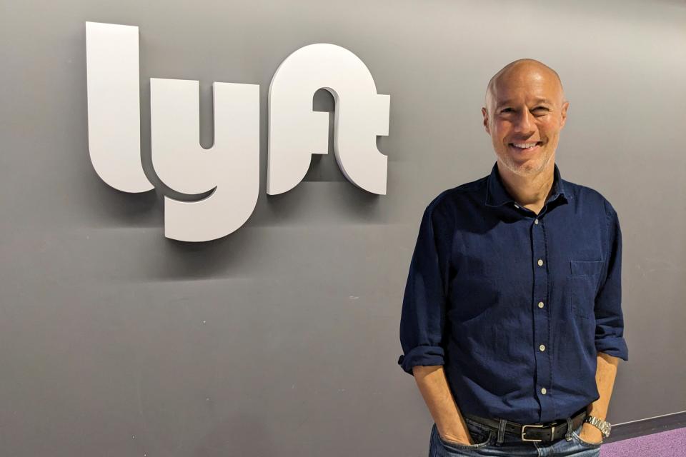 Lyft CEO David Risher standing in front of a gray wall with a white Lyft logo behind him.