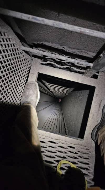 Two people at the top of a dark, narrow shaft with a metal grid floor, one person's shoes and legs partially visible