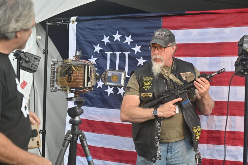 The Rod of Iron Freedom Festival 2021: A man holding an automatic rifle poses in front of a Three Percenter flag, which is a far-right anti-government militia movement.