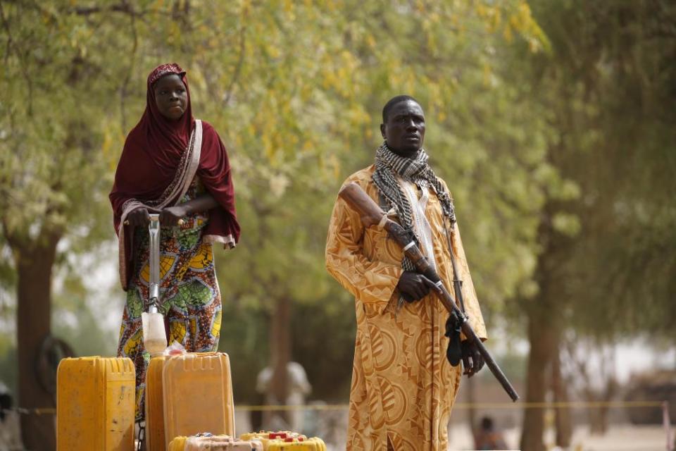 A man holds a hunting rifle as a woman pumps water into jerrycans in Kerawa, Cameroon