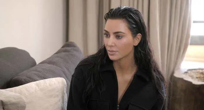 Kim Kardashian sits on a couch, wearing a dark zip-up top, with wet hair and a neutral expression, in a room with curtains in the background