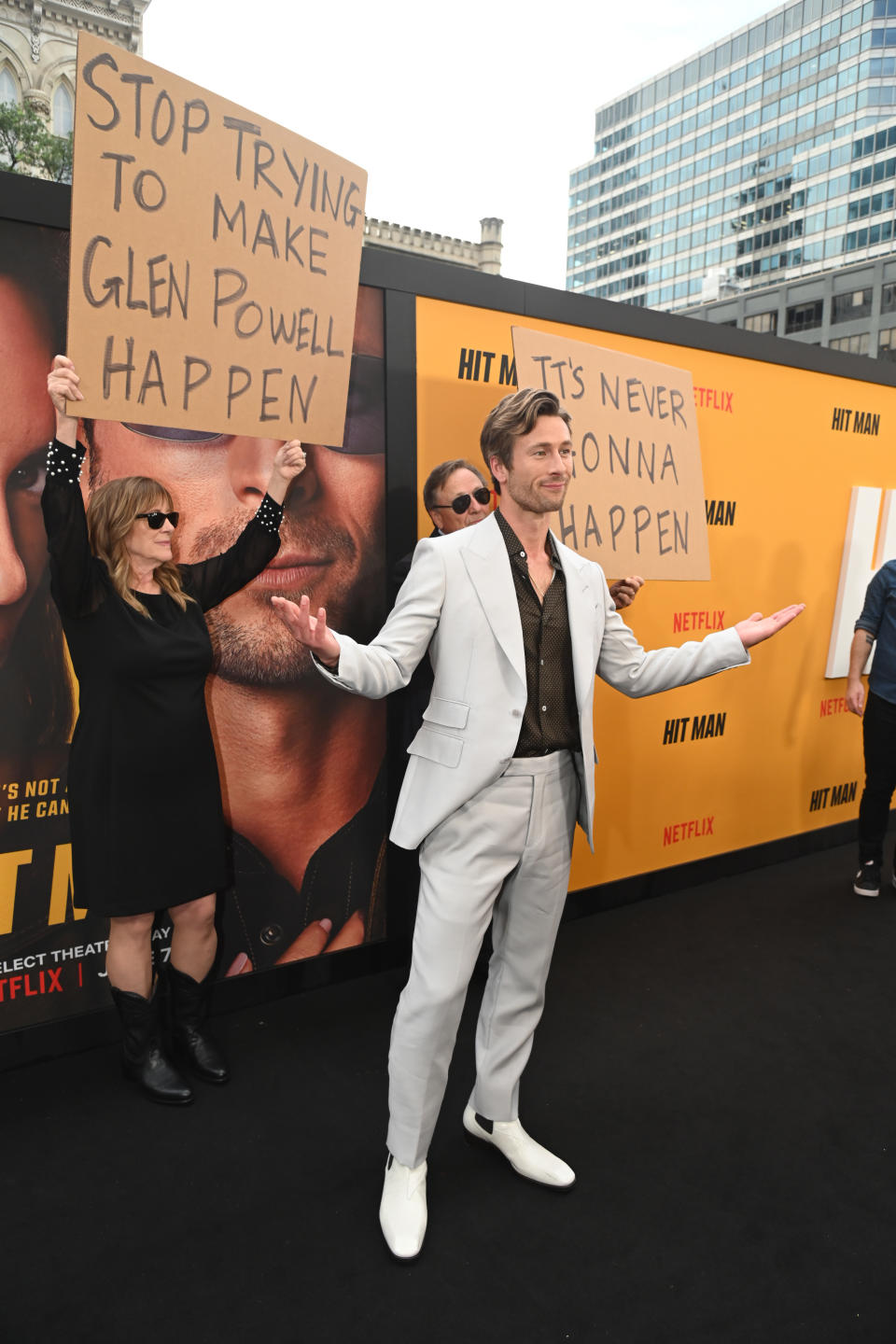 Glen Powell standing with a woman holding satirical protest signs at a movie premiere