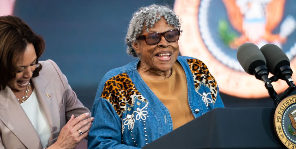 opal lee, wearing a denim designed coat, yellow shirt, and sunglasses, speaks at a podium with two microphones and the us presidential seal, standing next to a laughing kamala harris