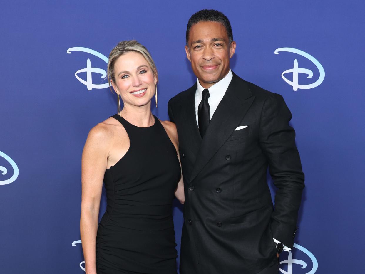 amy robach, wearing a blach dress with an asymmetric, ruffled hem, and t.j. holmes, wearing a black suit and black tie, standing together in front of a navy background with the Disney logo