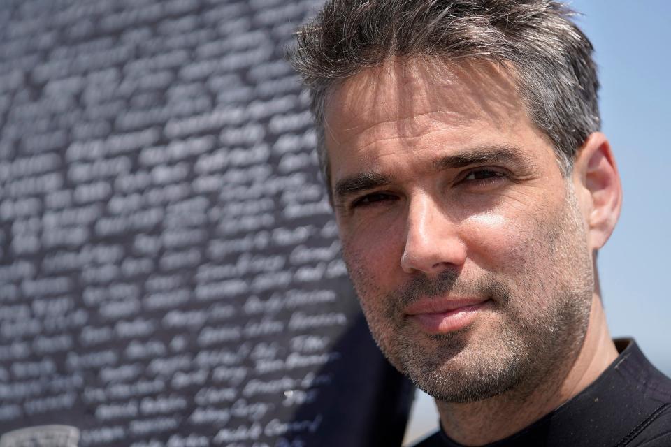 Dan Fischer, of Newport, R.I., stands for a photograph with his surfboard on Easton's Beach, in Newport, Wednesday, May 18, 2022. Fischer, 42, created the One Last Wave Project in January 2022 to use the healing power of the ocean to help families coping with a loss, as it helped him following the death of his father. Fischer places names of lost loved ones onto his surfboards, then takes the surfboards out into the ocean as a way to memorialize the loved ones in a place that was meaningful to them. (AP Photo/Steven Senne)