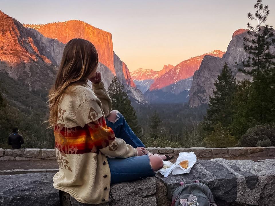 Emily sits on a wall and looks out at the mountains and trees at sunset. There is food next to her on the wall. Under the wall is a backpack covered in patches.