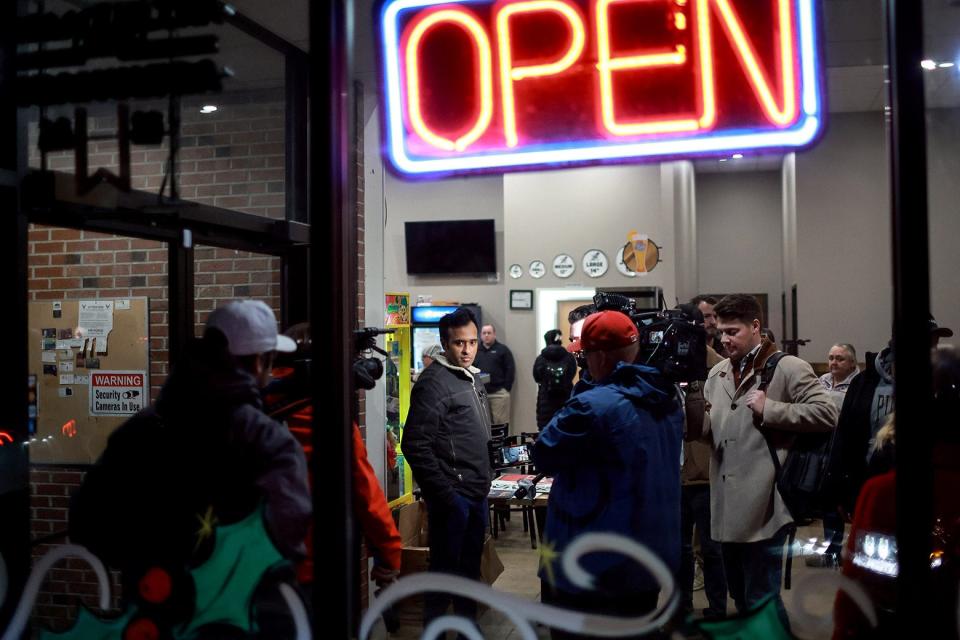 A man in line for pizza, wearing a parka, with a large, neon Open sign above his head.