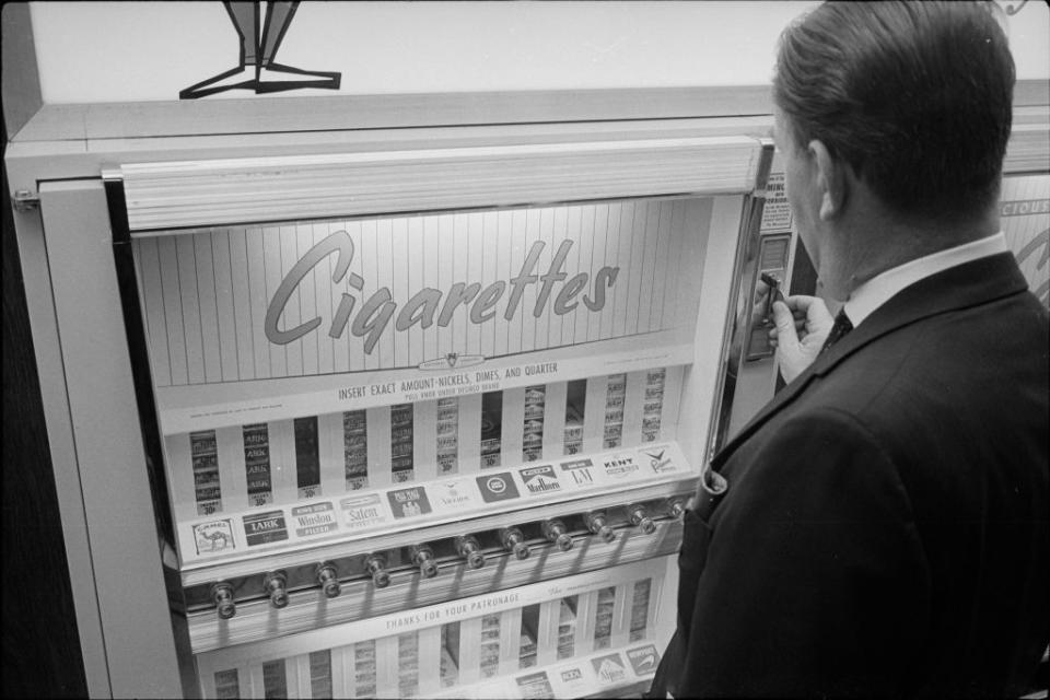 A man in a suit and tie using a cigarette vending machine labeled "Cigarettes." Various cigarette brands are visible in the machine