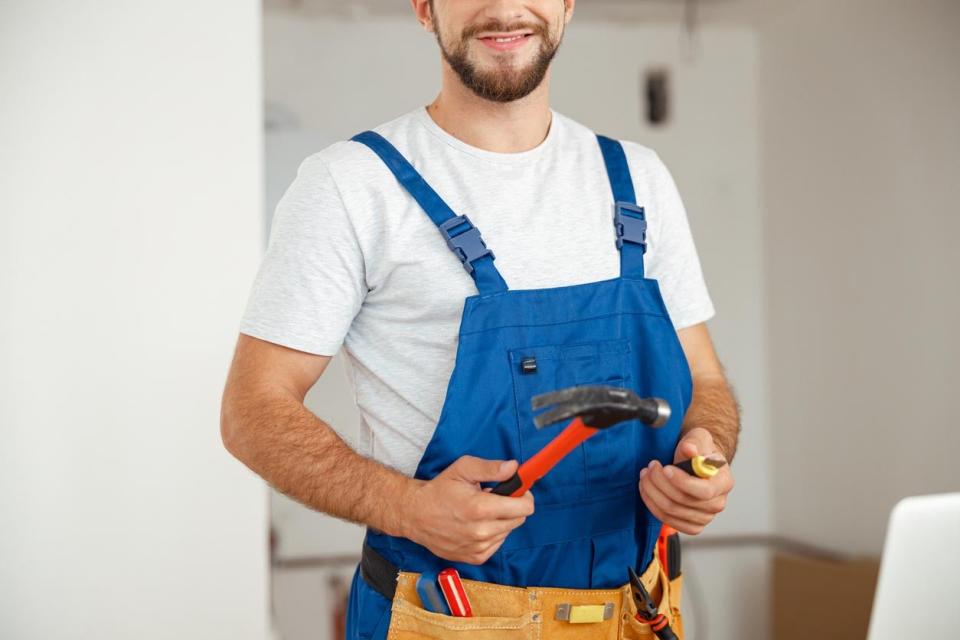A man in a blue apron, wearing a tool belt, holds a hammer.