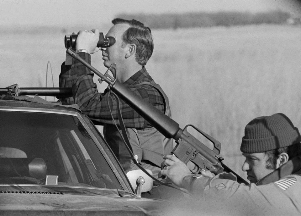 FILE - A U.S. marshal peers through binoculars as his partner keeps a weapon ready in the plains of Wounded Knee, S.D., on March 13, 1973, as government forces reinstated barricades along the roads leading to the village. (AP Photo/DT, File)