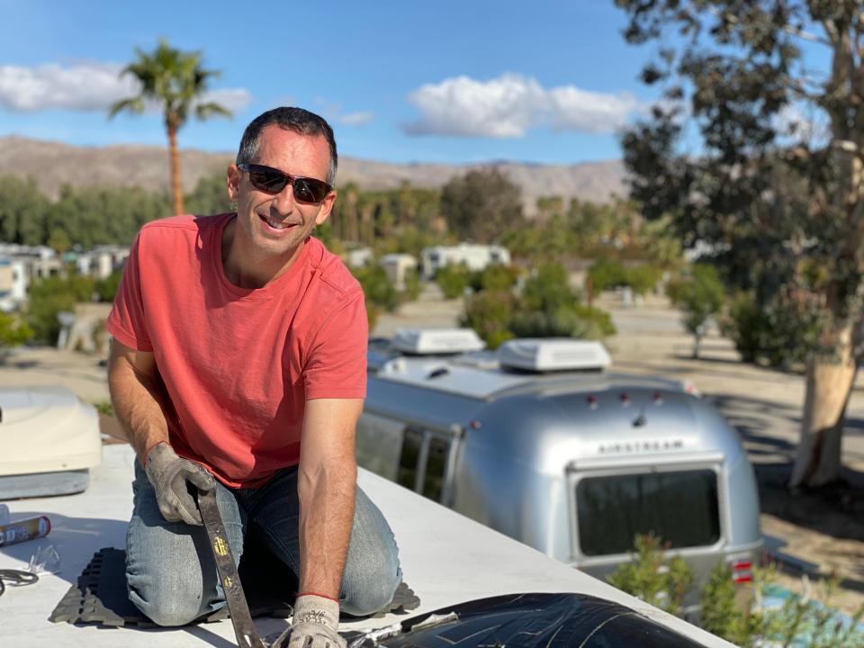 Man in sunglasses repairs an RV roof with an airstream and trees in the background. Blue skies.