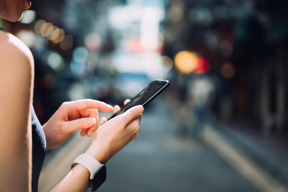 Close up and over the shoulder view of young Asian woman using smartphone while commuting in the city, against urban city scene