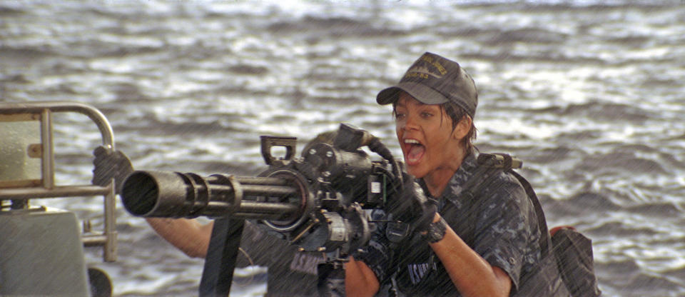 Rihanna in a naval uniform operates a large ship-mounted machine gun with an intense expression amidst a rainy backdrop