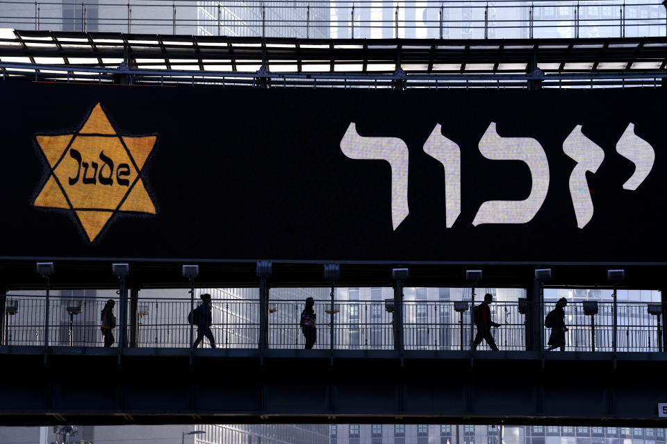 People walk along a bridge lit with a billboard showing a yellow Star of David that reads "Jude", Jew in German, resembling the one Jews were forced to wear in Nazi Germany and the word "remember"in Hebrew, during the annual Holocaust Remembrance Day in Ramat Gan, Israel, Tuesday, April 18, 2023. Israel marking the annual Day of Remembrance for the six million Jewish victims of the Nazi genocide who perished during World War II. (AP Photo/Oded Balilty)