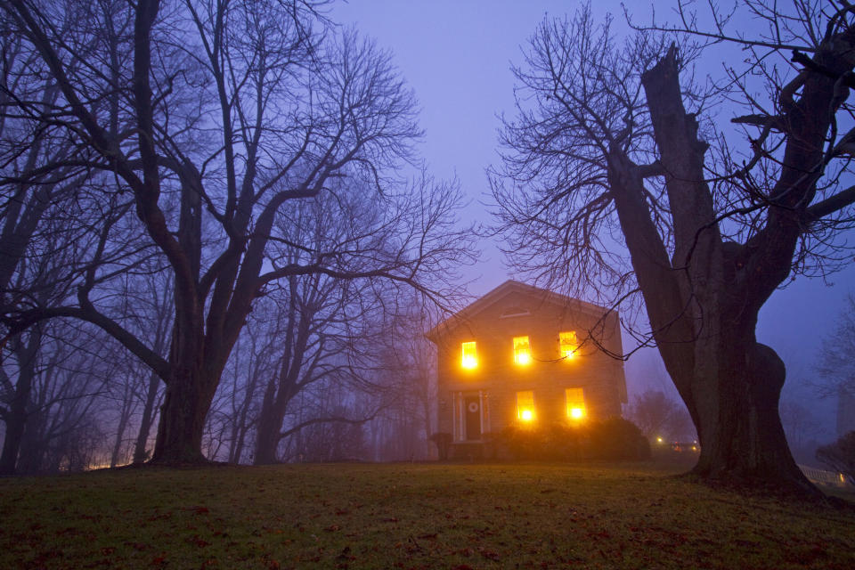 A misty evening scene showing a large, illuminated house surrounded by leafless trees. The house's warm lights glow against the foggy backdrop