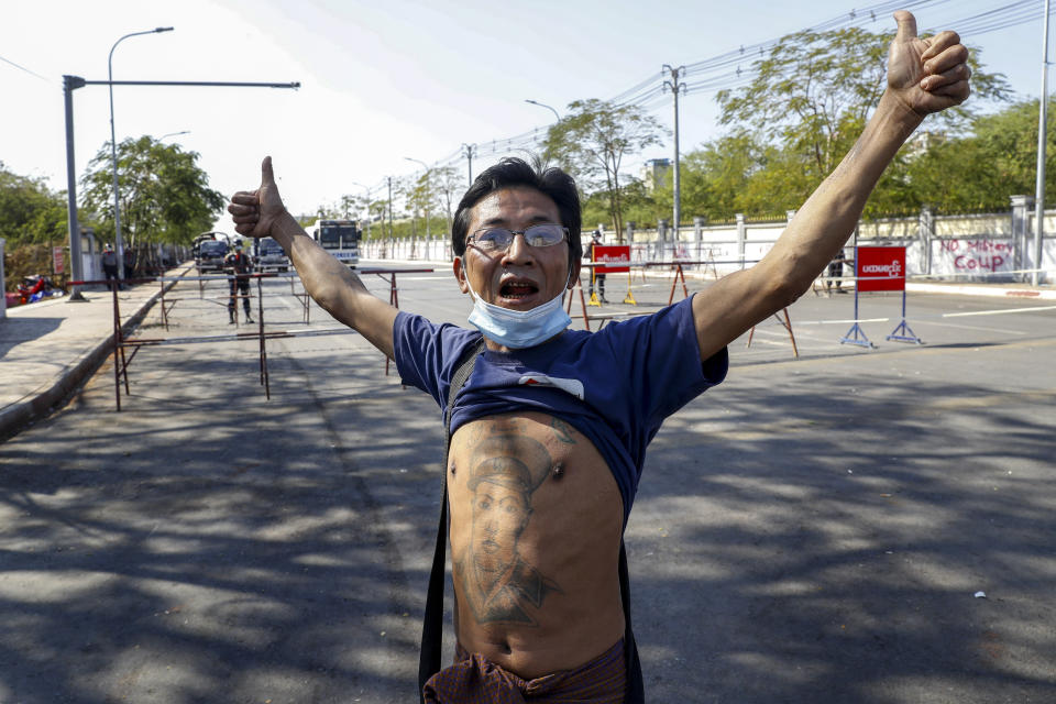 A man with a tattoo of Gen. Aung San, father of detained Myanmar leader Aung San Suu Kyi, gestures stands close to a cordoned-off road with police trucks and water cannons, the site of clashes between protesters and police a day earlier, in Mandalay, Myanmar, Wednesday, Feb. 10, 2021. Protesters continued to gather Wednesday in Mandalay breaching Myanmar's new military rulers' decrees that effectively banned peaceful public protests in the country's two biggest cities. (AP Photo)