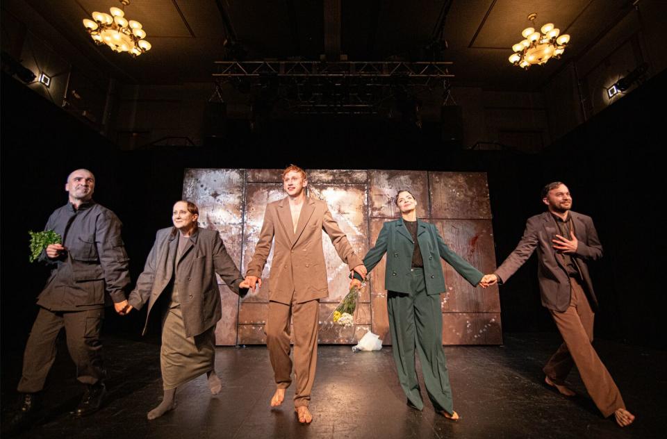 Five actors in loose-fitting suits hold hands on stage