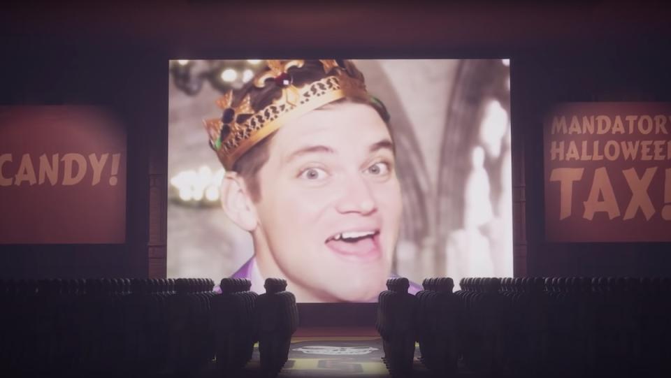 Nick Lutsko wears a crown on a big screen in front of an audience of bald male dolls in "Spirit Halloween Planet"
