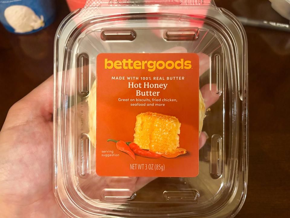 A hand holds a clear container of light-yellow butter with an orange label with "Bettergoods hot-honey butter" text on it
