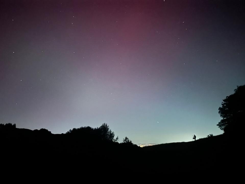 night sky with pink and blue glow above city lights peeking above the dark silhouette of a hill with a person on it looking out