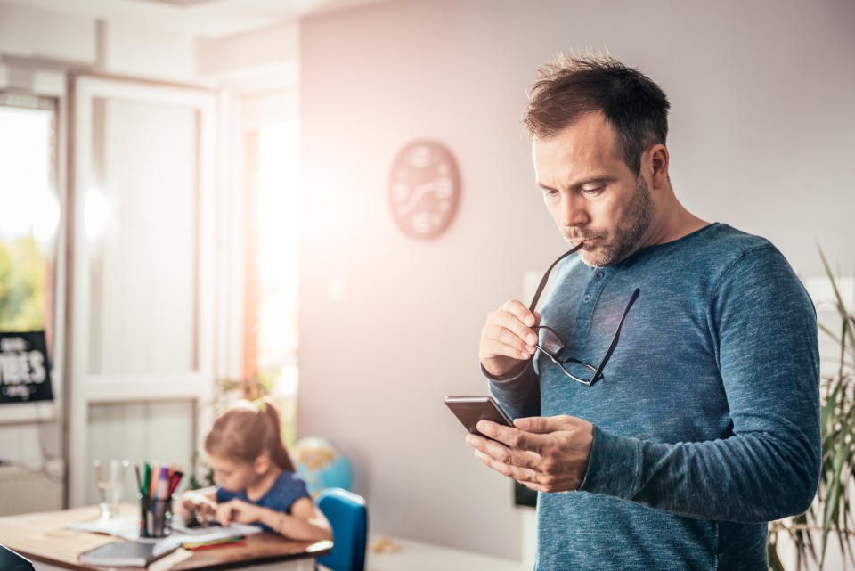 Worried father looking at smart phone with eyeglasses in his hand, in background his daughter doing homework. (Getty Images)