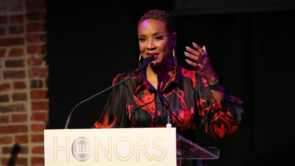 MC Lyte speaking at a podium in a black and red dress at the RIAA Honors ceremony